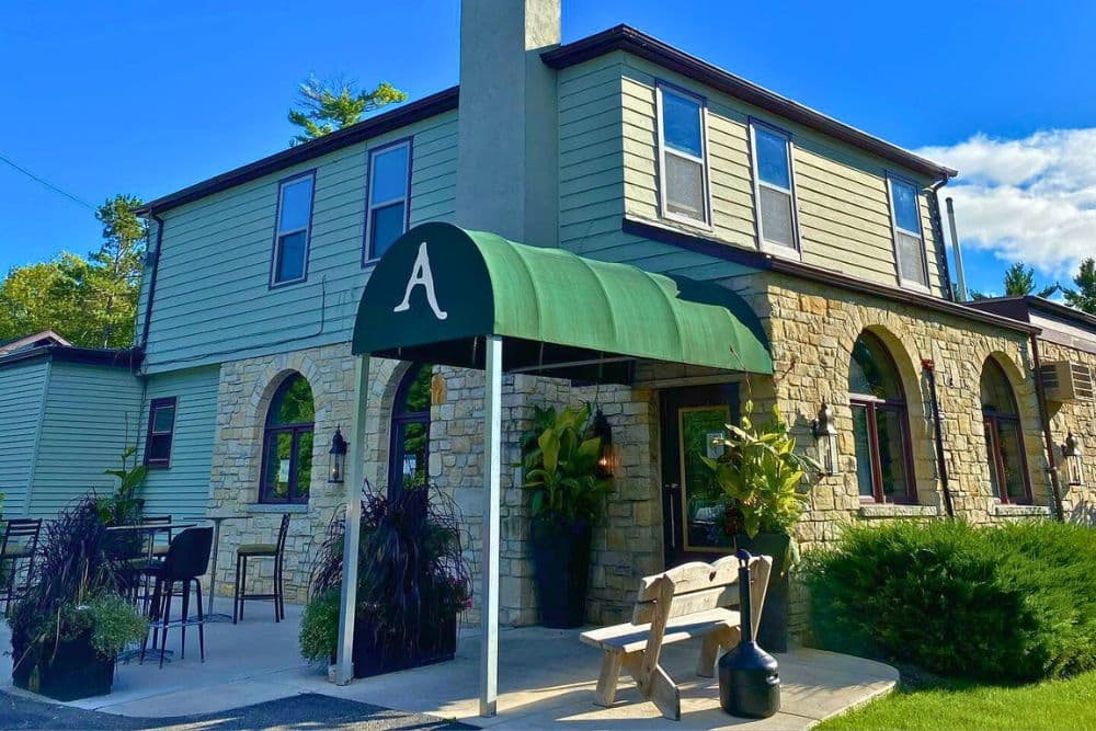 A green awning with a white "A" welcomes visitors to a two-story, stone and wood building with landscaped outdoor seating.