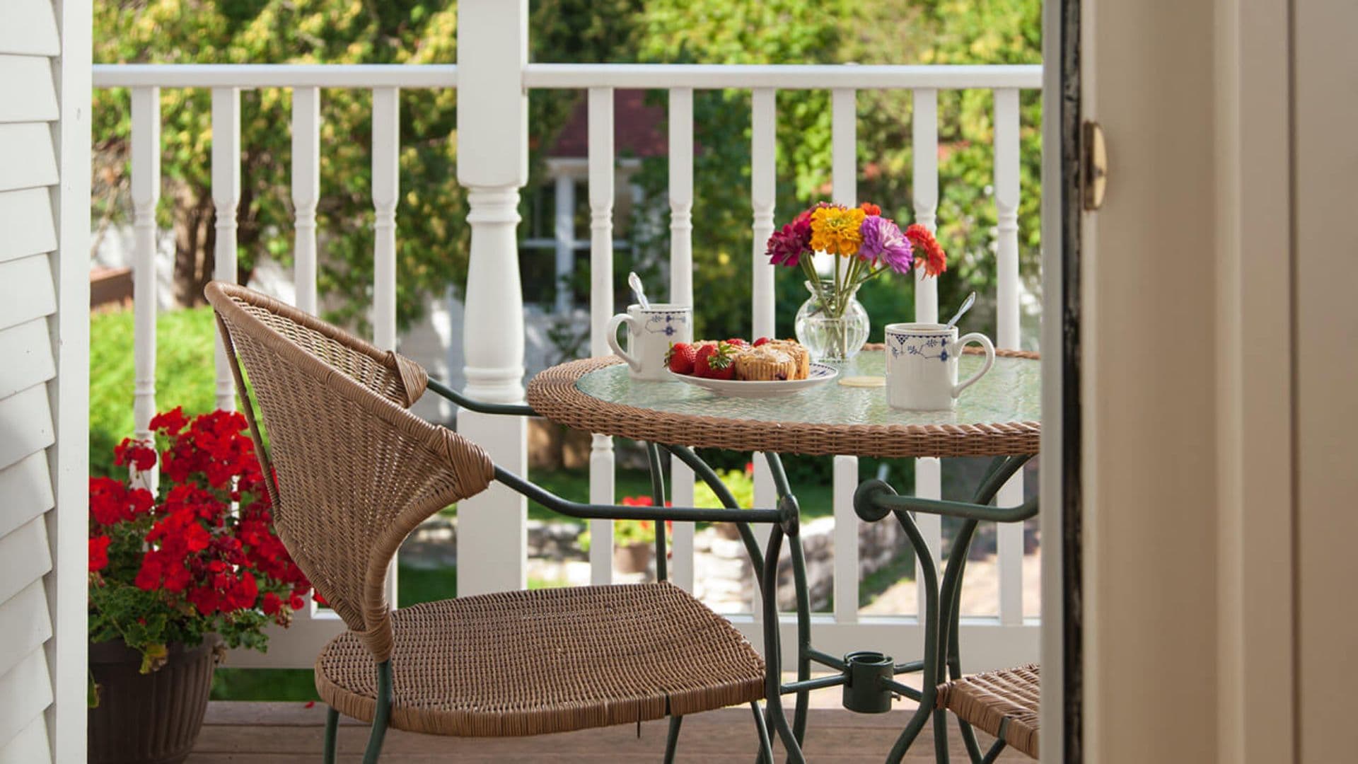 A cozy balcony with a table set for breakfast, featuring flowers, mugs, and pastries.