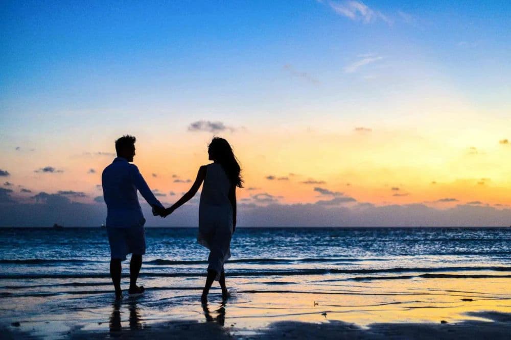 A couple walks hand in hand along a beach at sunset.