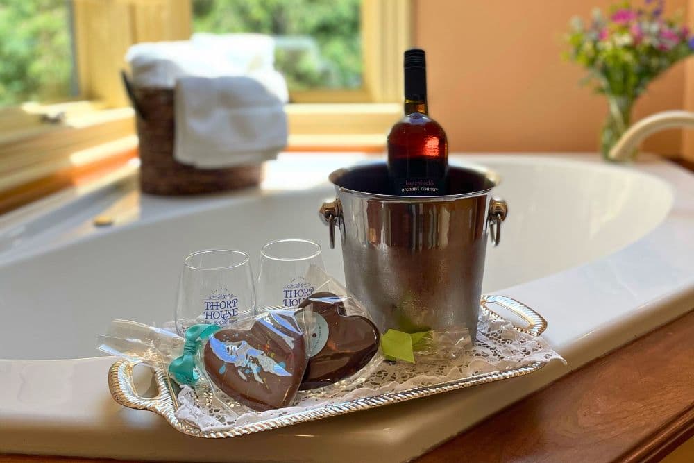 A silver tray holds two glasses, chocolate hearts, and a wine bottle, set beside a luxurious bathtub.